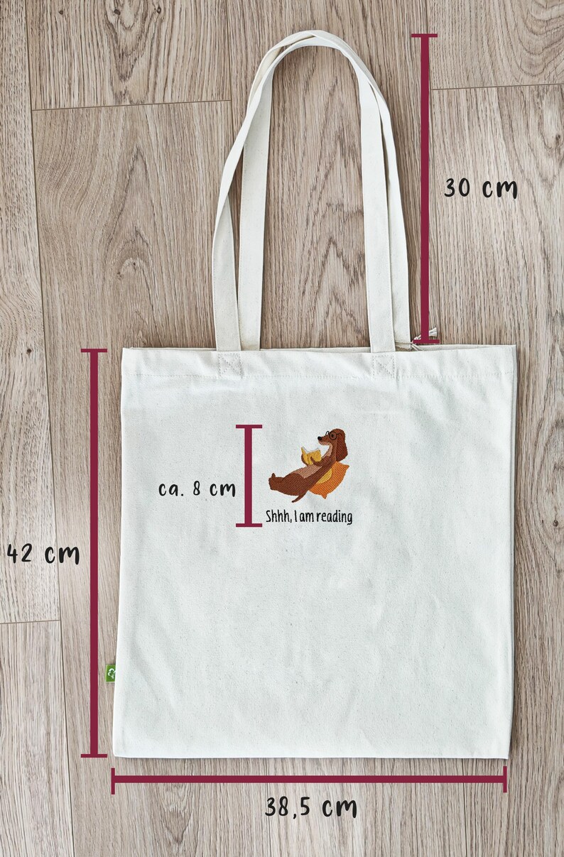 Puede incluir: Una bolsa de tela blanca con un dachshund leyendo un libro. La bolsa tiene asas marrones y mide 38,5 cm de ancho, 42 cm de alto y 30 cm de profundidad. El texto en la bolsa dice "Shhh, I am reading."