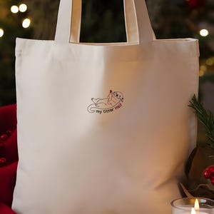 Puede incluir: Un bolso de tela color crema con un bordado que muestra una nutria tumbada boca arriba sosteniendo un coraz&oacute;n rojo. Debajo de la nutria, el texto "my Otter Half" aparece en letras negras. El bolso cuenta con dos asas de tela a juego.