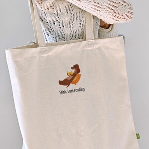 Puede incluir: Una bolsa de lona blanca con un perro salchicha marr&oacute;n leyendo un libro y el texto "Shhh... I am reading".