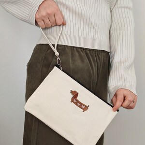 May include: A white canvas zippered pouch with a black trim and a brown dachshund with pink hearts embroidered on the front.