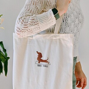Puede incluir: Una bolsa de lona blanca con un dachshund marr&oacute;n haciendo yoga en una colchoneta. El texto "100% YOGI" est&aacute; bordado debajo del dachshund.