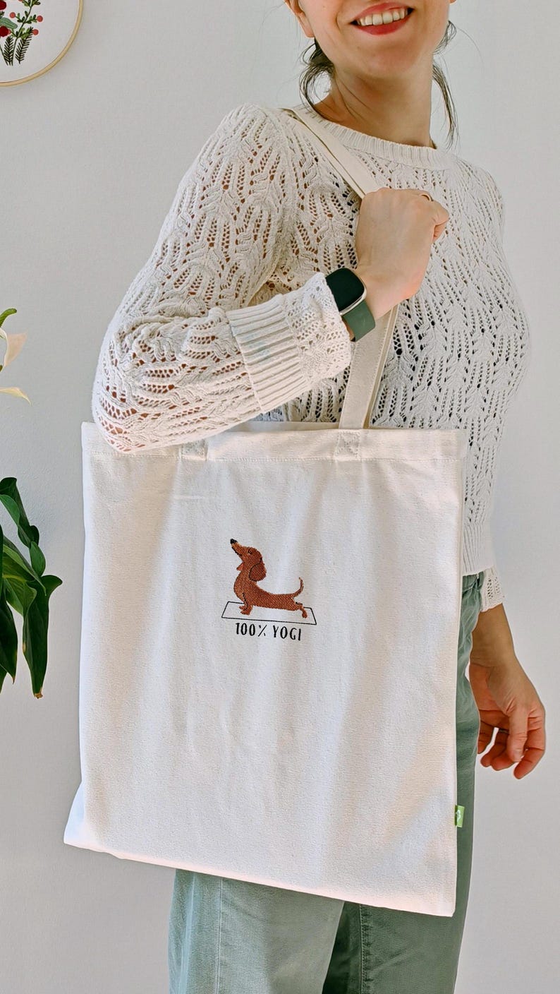 Puede incluir: Una bolsa de lona blanca con un dachshund marr&oacute;n haciendo yoga en una colchoneta. El texto "100% YOGI" est&aacute; bordado debajo del dachshund.