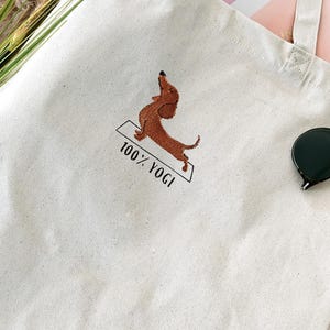 Puede incluir: Una bolsa de lona blanca con un dachshund marr&oacute;n bordado en ella haciendo una pose de yoga. El texto "100% YOGI" est&aacute; bordado debajo del dachshund.