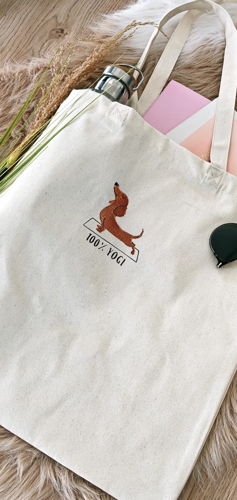Puede incluir: Una bolsa de lona blanca con un dachshund marr&oacute;n bordado en ella haciendo una pose de yoga. El texto "100% YOGI" est&aacute; bordado debajo del dachshund.