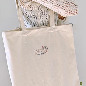 Puede incluir: Un bolso de tela color crema con un bordado de una nutria marina que sostiene un peque&ntilde;o coraz&oacute;n rojo. El texto "my Otter Half" est&aacute; cosido en negro debajo de la nutria. El bolso tiene dos asas largas para el hombro y una etiqueta verde.