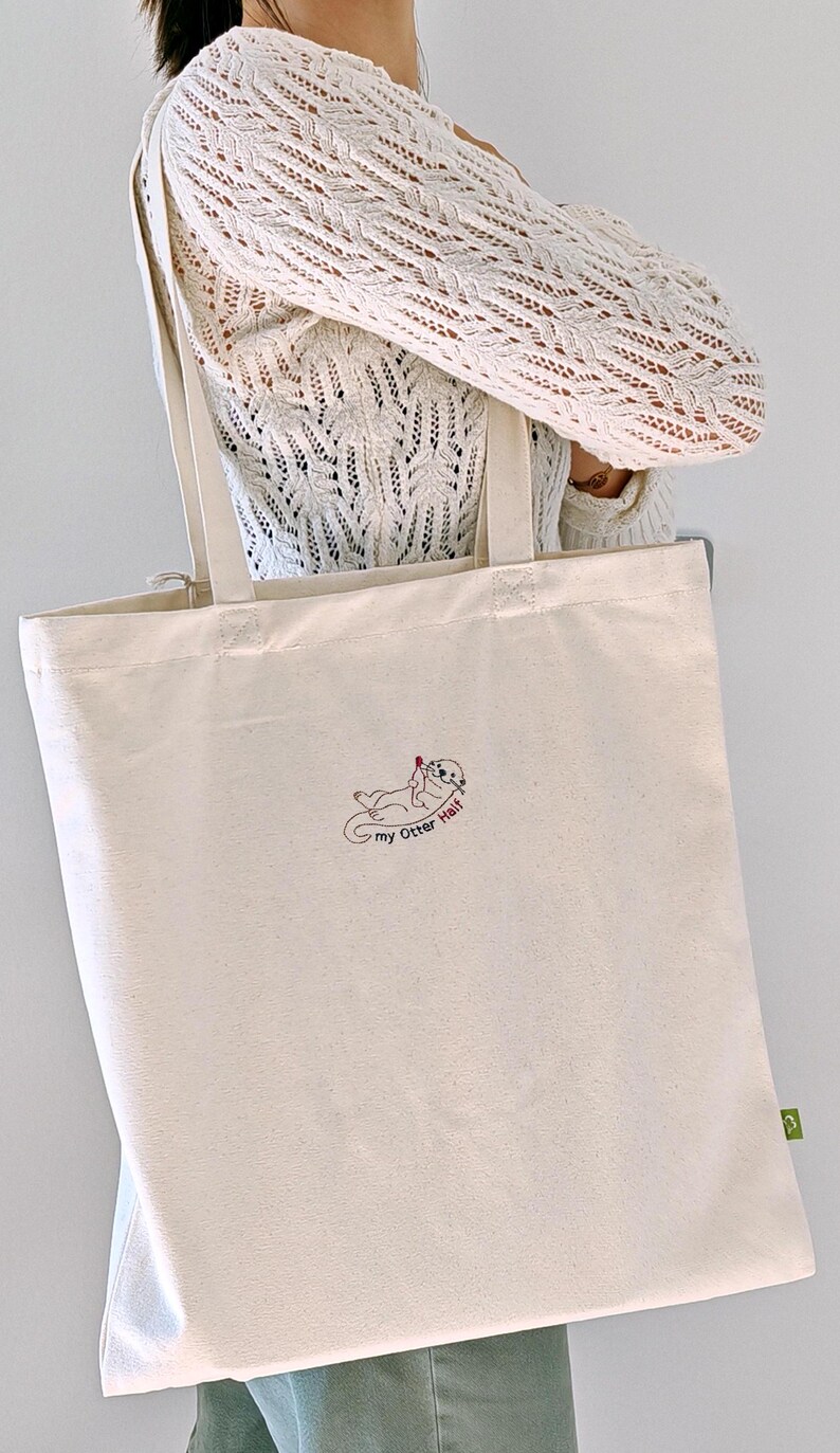Puede incluir: Un bolso de tela color crema con un bordado de una nutria marina que sostiene un peque&ntilde;o coraz&oacute;n rojo. El texto "my Otter Half" est&aacute; cosido en negro debajo de la nutria. El bolso tiene dos asas largas para el hombro y una etiqueta verde.