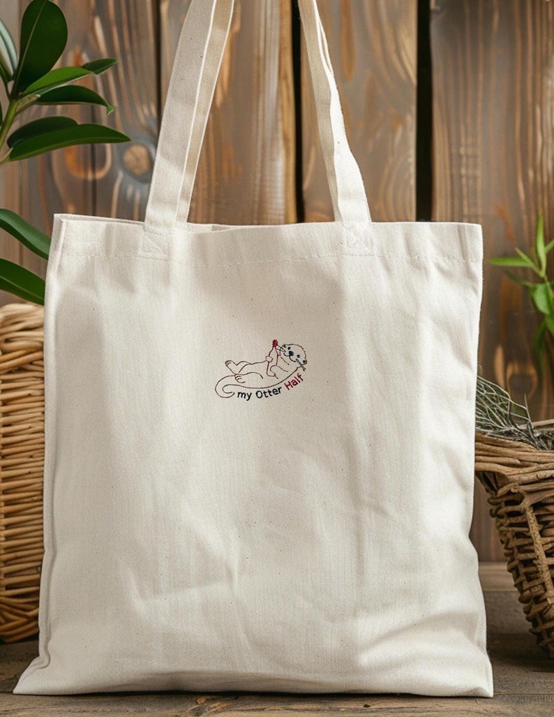 Puede incluir: Un bolso de tela color crema con una ilustraci&oacute;n bordada de una nutria marina tumbada boca arriba sosteniendo un peque&ntilde;o coraz&oacute;n rojo. Debajo de la nutria, el texto "my Otter Half" aparece en letras bordadas de color negro y rojo.