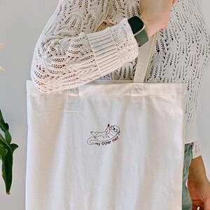 Puede incluir: Un bolso de tela color crema con un peque&ntilde;o bordado de una nutria tumbada boca arriba sosteniendo un coraz&oacute;n rojo. Debajo de la nutria, el texto "my Otter Half" est&aacute; bordado en letras cursivas de color marr&oacute;n oscuro.