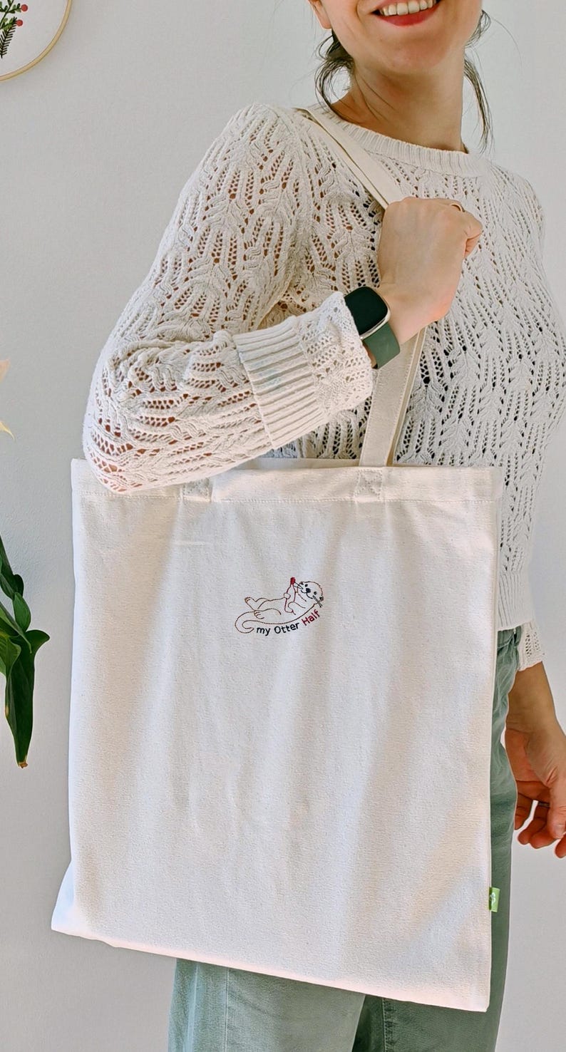 Puede incluir: Un bolso de tela color crema con un peque&ntilde;o bordado de una nutria tumbada boca arriba sosteniendo un coraz&oacute;n rojo. Debajo de la nutria, el texto "my Otter Half" est&aacute; bordado en letras cursivas de color marr&oacute;n oscuro.