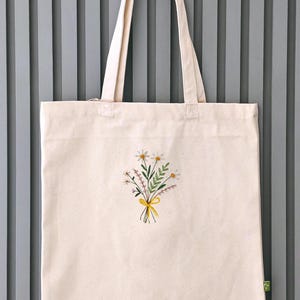 May include: A white canvas tote bag with a small embroidered bouquet of flowers in the center. The flowers are yellow, white, and green. The bag has two long straps.