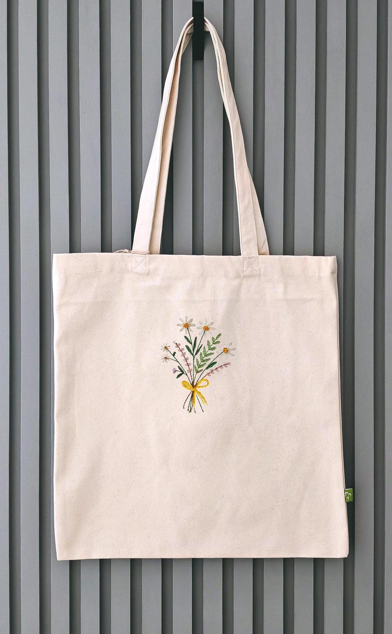 May include: A white canvas tote bag with a small embroidered bouquet of flowers in the center. The flowers are yellow, white, and green. The bag has two long straps.