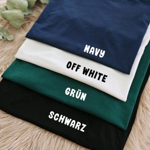 May include: A stack of folded t-shirts in navy blue, off-white, dark green, and black. Each shirt has a label with its color name in white block letters. The shirts are arranged on a light brown, textured surface.