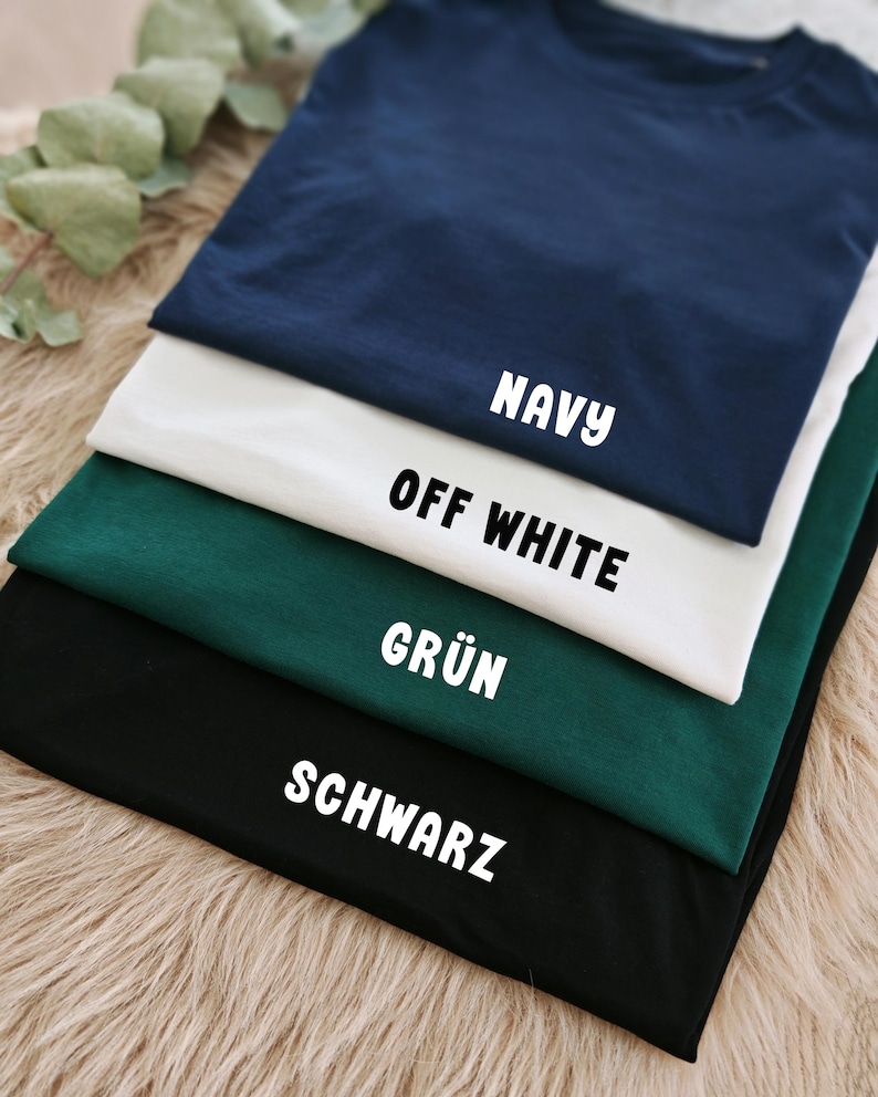 May include: A stack of folded t-shirts in navy blue, off-white, dark green, and black. Each shirt has a label with its color name in white block letters. The shirts are arranged on a light brown, textured surface.