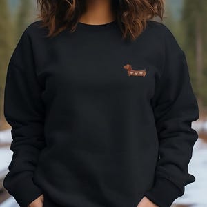 May include: Black sweatshirt featuring a small embroidered dachshund with heart-shaped markings. The dog is brown with white and pink hearts. The sweatshirt has a crew neck and long sleeves. The model is wearing blue jeans.
