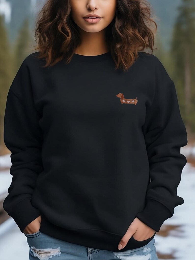 May include: Black sweatshirt featuring a small embroidered dachshund with heart-shaped markings. The dog is brown with white and pink hearts. The sweatshirt has a crew neck and long sleeves. The model is wearing blue jeans.