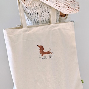 Puede incluir: Una bolsa de lona beige con un perro salchicha marr&oacute;n bordado haciendo yoga. El texto "100% YOGI" est&aacute; bordado debajo del perro.