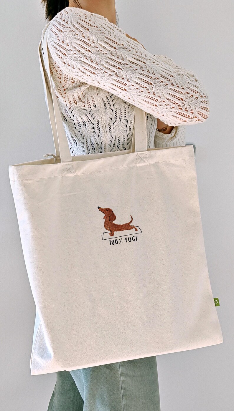 Puede incluir: Una bolsa de lona beige con un perro salchicha marr&oacute;n bordado haciendo yoga. El texto "100% YOGI" est&aacute; bordado debajo del perro.