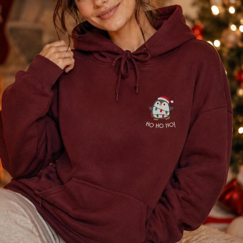 May include: A maroon hooded sweatshirt featuring a drawstring hood and a front kangaroo pocket. An embroidered graphic on the chest depicts a penguin wearing a Santa hat and wrapped in colorful Christmas lights, with the text HO HO HO below.