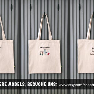 Puede incluir: Tres bolsas de tela de lona blanca con diferentes dise&ntilde;os bordados. La primera bolsa dice "Merry Woofmas" y presenta un dachshund. La segunda bolsa dice "Merry Catmas" y presenta un gato con adornos. La tercera bolsa dice "Winter Love" y presenta un ping&uuml;ino.