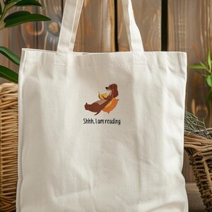 Puede incluir: Una bolsa de lona blanca con un dachshund marr&oacute;n que lleva gafas y sostiene un pl&aacute;tano. El texto "Shhh, I am reading" est&aacute; bordado en la bolsa.