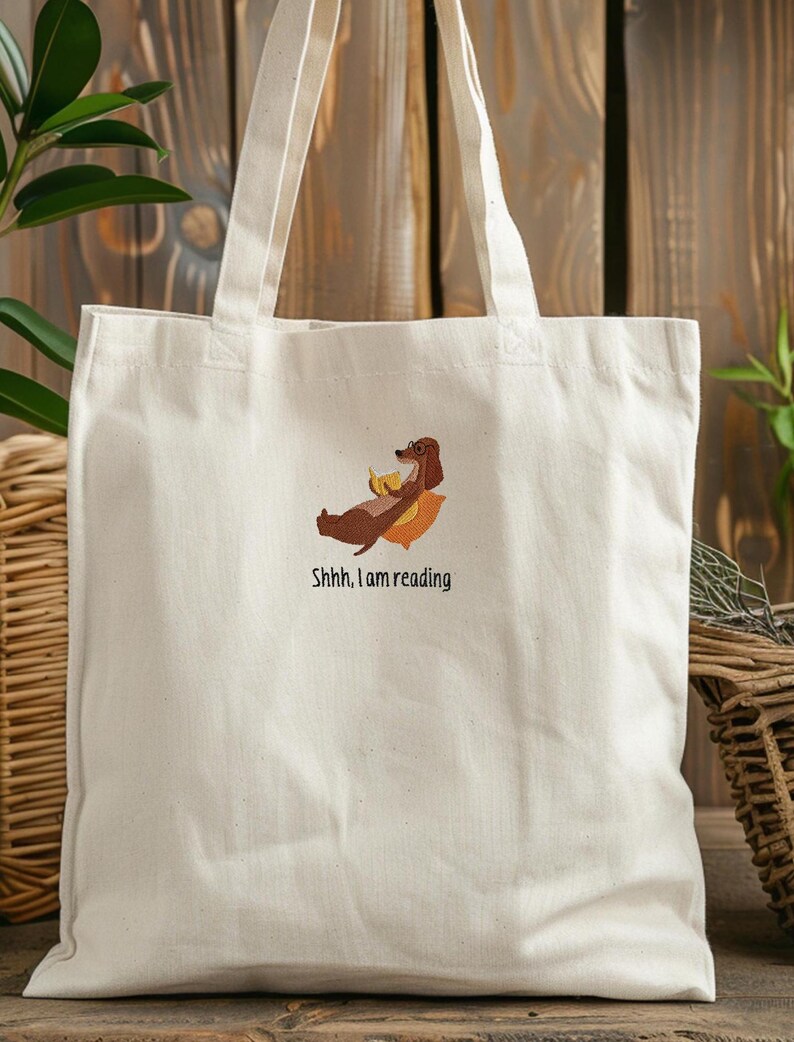 Puede incluir: Una bolsa de lona blanca con un dachshund marr&oacute;n que lleva gafas y sostiene un pl&aacute;tano. El texto "Shhh, I am reading" est&aacute; bordado en la bolsa.