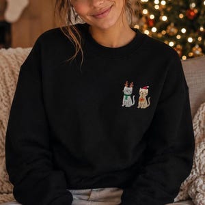 May include: A black crewneck sweatshirt featuring an embroidered design on the left chest. The embroidery depicts two cats: one grey cat wearing brown reindeer antlers and a green scarf, and one tan cat wearing a red and white Santa hat.