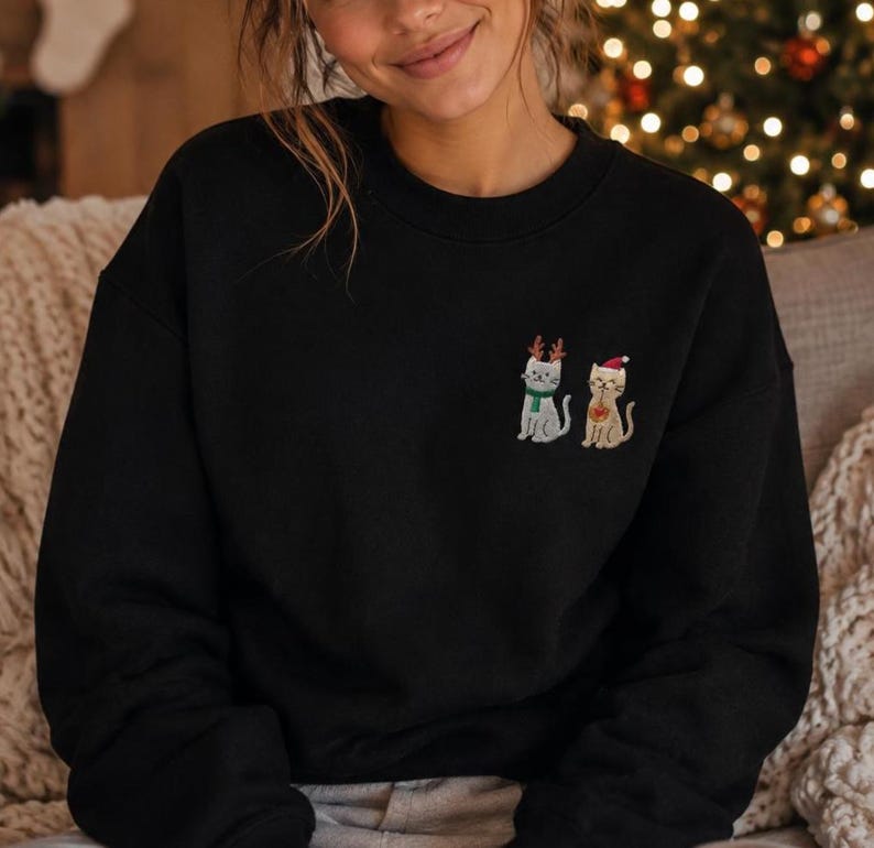 May include: A black crewneck sweatshirt featuring an embroidered design on the left chest. The embroidery depicts two cats: one grey cat wearing brown reindeer antlers and a green scarf, and one tan cat wearing a red and white Santa hat.
