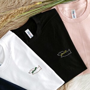 May include: Three t-shirts with different colored backgrounds and a dachshund design embroidered on each. The t-shirts are white, black, and pink. The dachshund is wearing a green scarf. The text "JERZEES" is visible on the pink t-shirt.