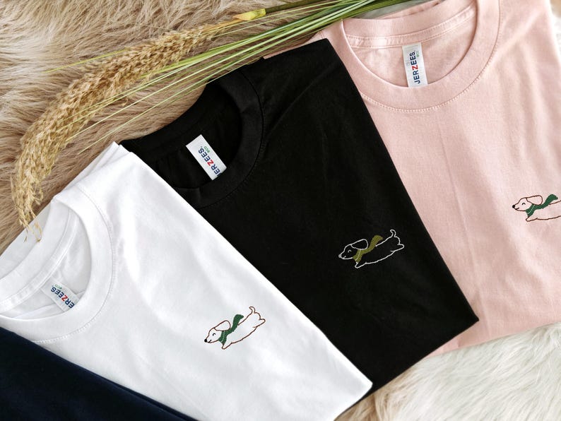 May include: Three t-shirts with different colored backgrounds and a dachshund design embroidered on each. The t-shirts are white, black, and pink. The dachshund is wearing a green scarf. The text "JERZEES" is visible on the pink t-shirt.