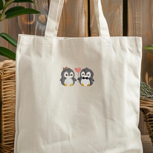 Puede incluir: Un bolso de tela de color natural con un dise&ntilde;o bordado de dos ping&uuml;inos. Uno lleva un peque&ntilde;o lazo marr&oacute;n y el otro una pajarita negra. Entre ambos, un peque&ntilde;o coraz&oacute;n rosa se sostiene sobre un tallo fino, centrado en el bolso.