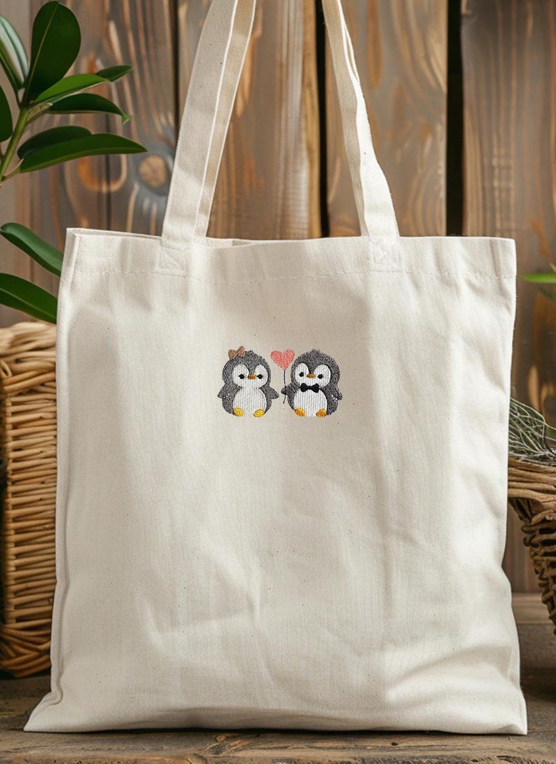 Puede incluir: Un bolso de tela de color natural con un dise&ntilde;o bordado de dos ping&uuml;inos. Uno lleva un peque&ntilde;o lazo marr&oacute;n y el otro una pajarita negra. Entre ambos, un peque&ntilde;o coraz&oacute;n rosa se sostiene sobre un tallo fino, centrado en el bolso.