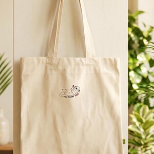 Puede incluir: Un bolso de tela color crema con dos asas largas para el hombro. El centro del bolso presenta un peque&ntilde;o gr&aacute;fico bordado de una nutria marina tumbada boca arriba sosteniendo un coraz&oacute;n rojo, con el texto "my Otter Half" escrito debajo.
