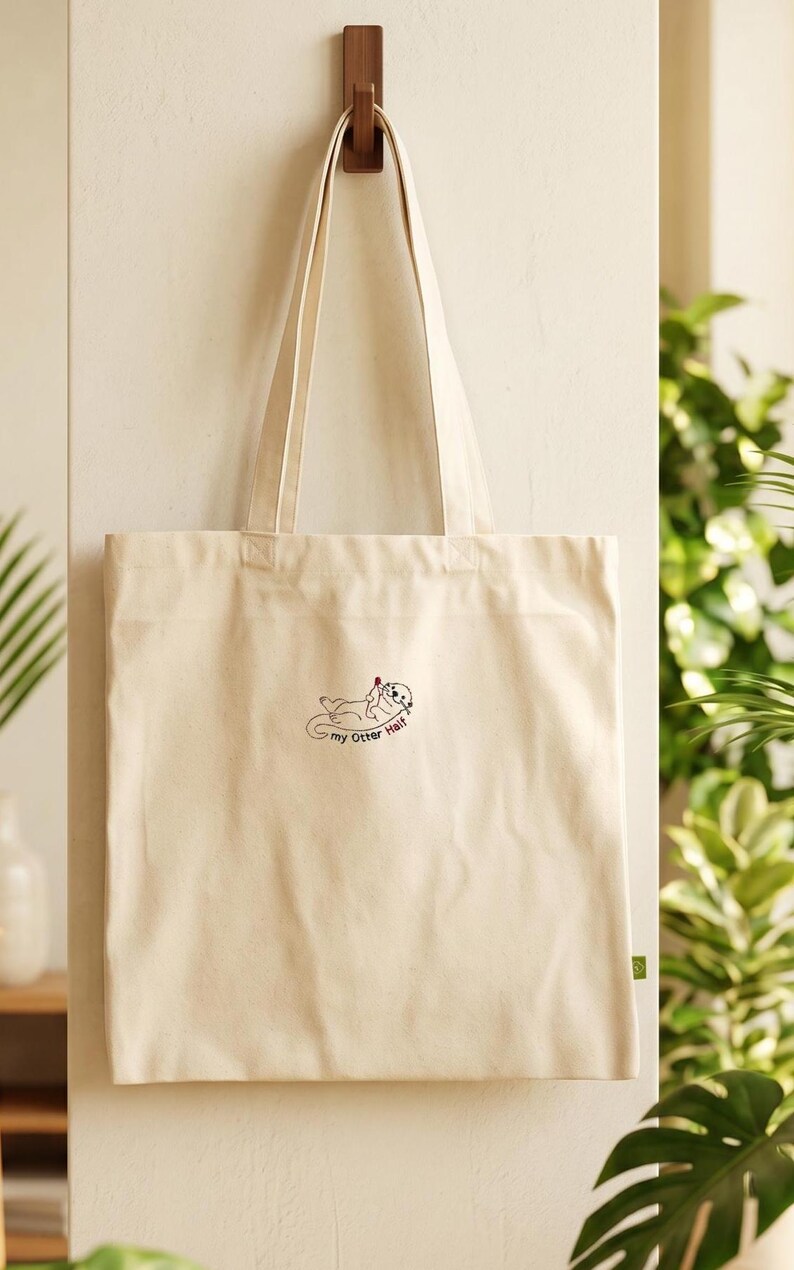 Puede incluir: Un bolso de tela color crema con dos asas largas para el hombro. El centro del bolso presenta un peque&ntilde;o gr&aacute;fico bordado de una nutria marina tumbada boca arriba sosteniendo un coraz&oacute;n rojo, con el texto "my Otter Half" escrito debajo.