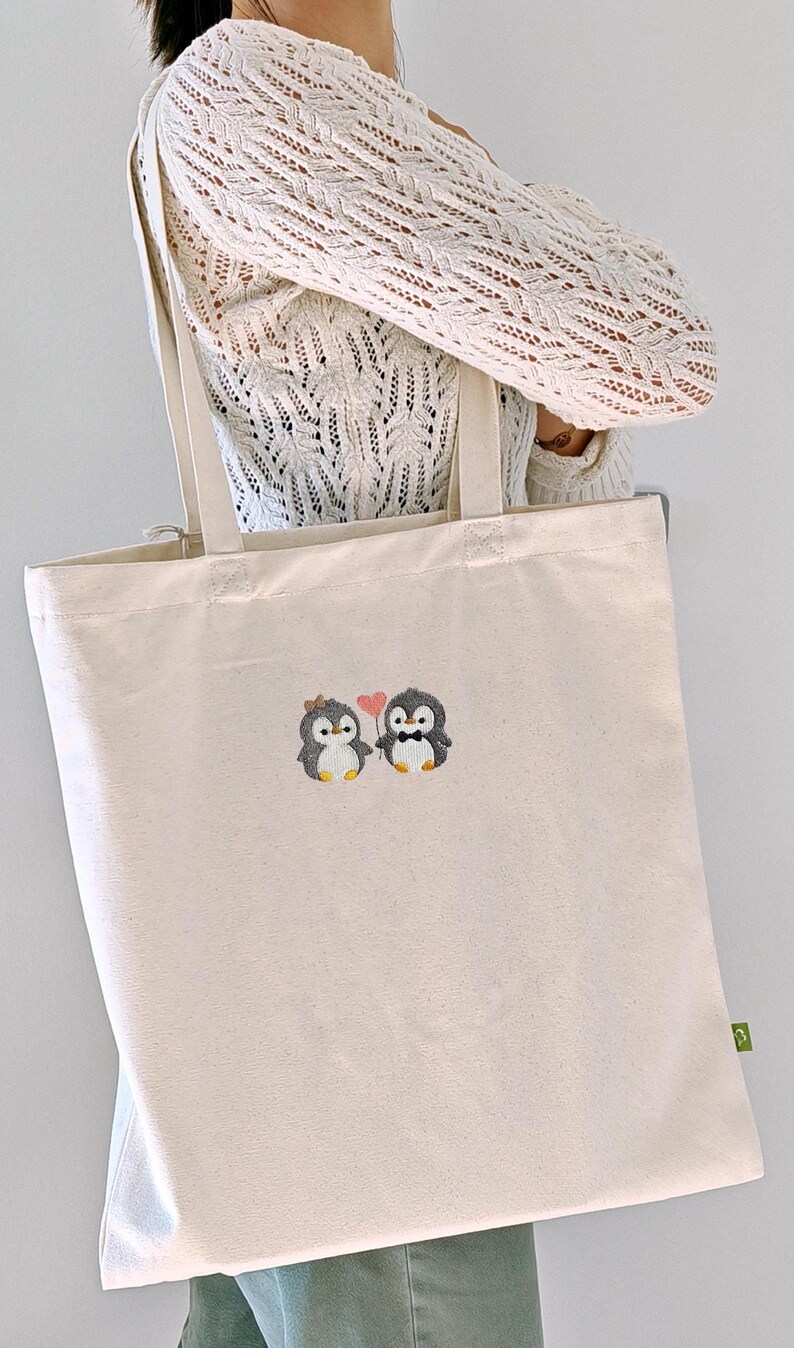 Puede incluir: Un bolso de tela de color natural con un dise&ntilde;o bordado de dos peque&ntilde;os ping&uuml;inos. Un ping&uuml;ino lleva un peque&ntilde;o lazo en la cabeza y el otro una pajarita negra. Entre ambos, sostienen un globo rosa en forma de coraz&oacute;n sujeto por una cuerda.
