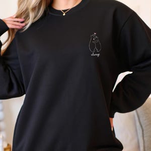 Op de afbeelding: Een zwart sweatshirt met lange mouwen en een ronde hals, voorzien van een kleine witte lijntekening op de linkerborst. De afbeelding toont twee beren die elkaar omhelzen met een klein roze hartje erboven en het woord always in wit cursief.