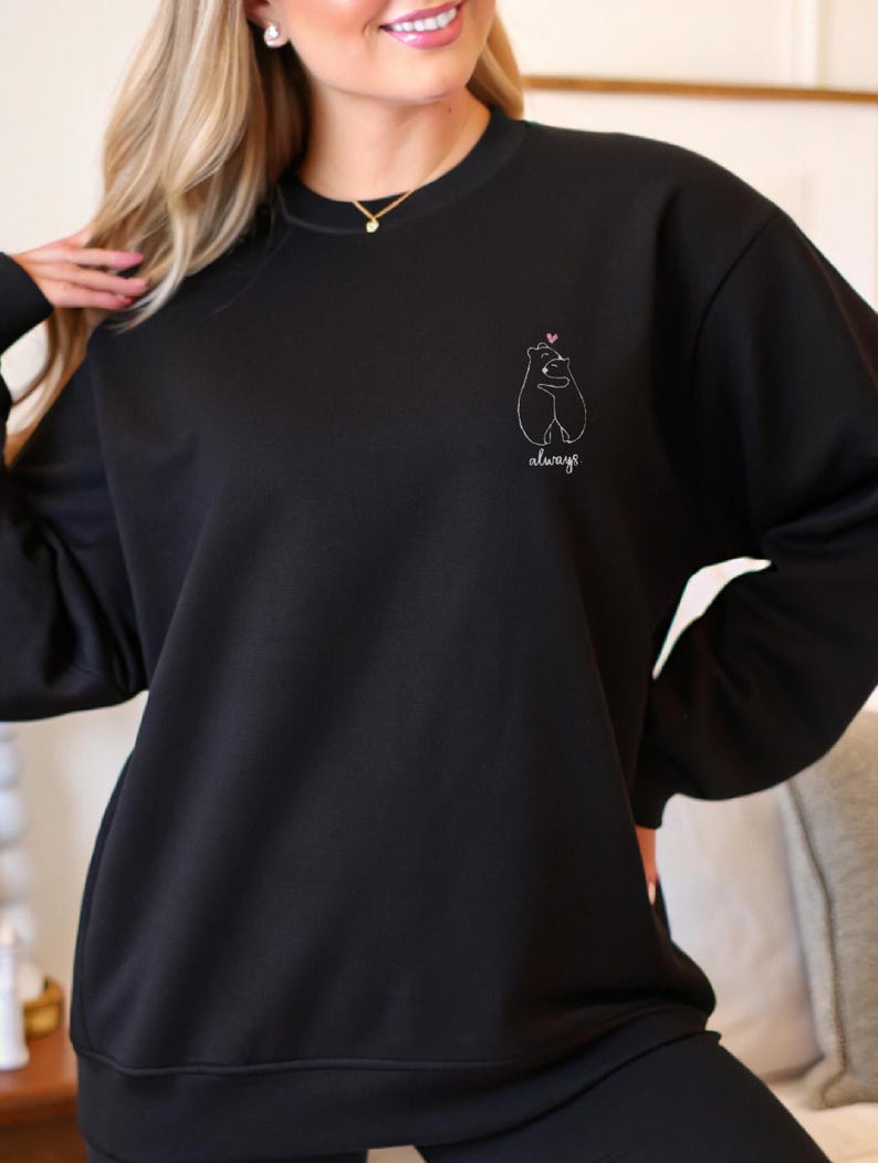 Op de afbeelding: Een zwart sweatshirt met lange mouwen en een ronde hals, voorzien van een kleine witte lijntekening op de linkerborst. De afbeelding toont twee beren die elkaar omhelzen met een klein roze hartje erboven en het woord always in wit cursief.