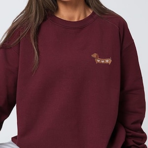 May include: A maroon sweatshirt with a small embroidered dachshund dog design on the chest. The dog is brown with white heart accents. The sweatshirt has a crew neck and long sleeves.