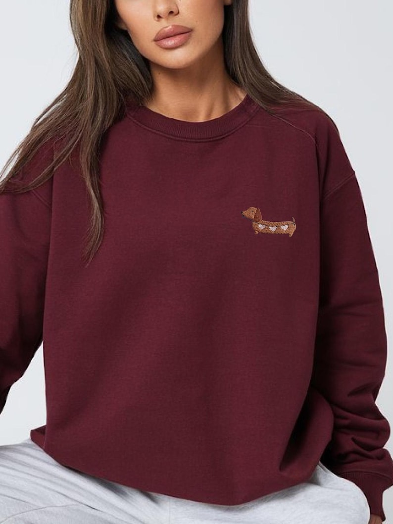 May include: A maroon sweatshirt with a small embroidered dachshund dog design on the chest. The dog is brown with white heart accents. The sweatshirt has a crew neck and long sleeves.
