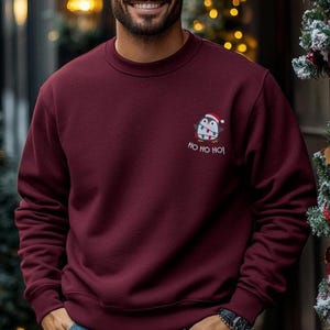 Mann trägt einen Pinguin Weihnachts Hoodie mit kleiner Stickerei vor weihnachtlichem Hintergrund.