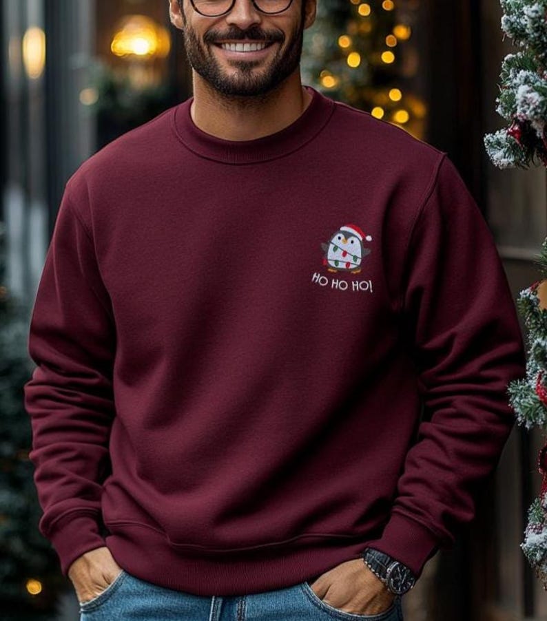 Mann trägt einen Pinguin Weihnachts Hoodie mit kleiner Stickerei vor weihnachtlichem Hintergrund.