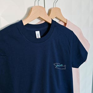 May include: Two t-shirts hanging on wooden hangers. The shirt on the left is navy blue with a white outline of a dachshund wearing a scarf. The shirt on the right is pink.