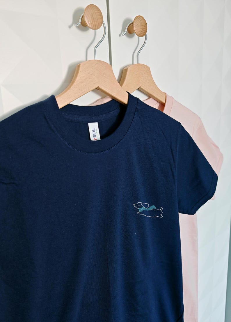 May include: Two t-shirts hanging on wooden hangers. The shirt on the left is navy blue with a white outline of a dachshund wearing a scarf. The shirt on the right is pink.
