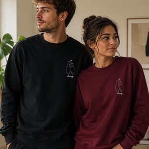 Op de afbeelding: Een zwarte sweater met ronde hals en een bordeauxrode sweater met ronde hals, beide voorzien van een kleine witte lijntekening op de linkerborst met twee knuffelende beren, een klein roze hartje en het woord always eronder geschreven.