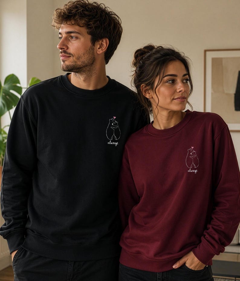 Op de afbeelding: Een zwarte sweater met ronde hals en een bordeauxrode sweater met ronde hals, beide voorzien van een kleine witte lijntekening op de linkerborst met twee knuffelende beren, een klein roze hartje en het woord always eronder geschreven.