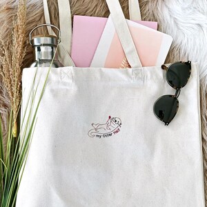 Puede incluir: Un bolso de tela color crema con un bordado de una nutria sosteniendo un coraz&oacute;n rojo y el texto "my Otter Half". El bolso contiene una botella de agua de metal plateado, papeles de color rosa y blanco, y tallos de hierba seca.