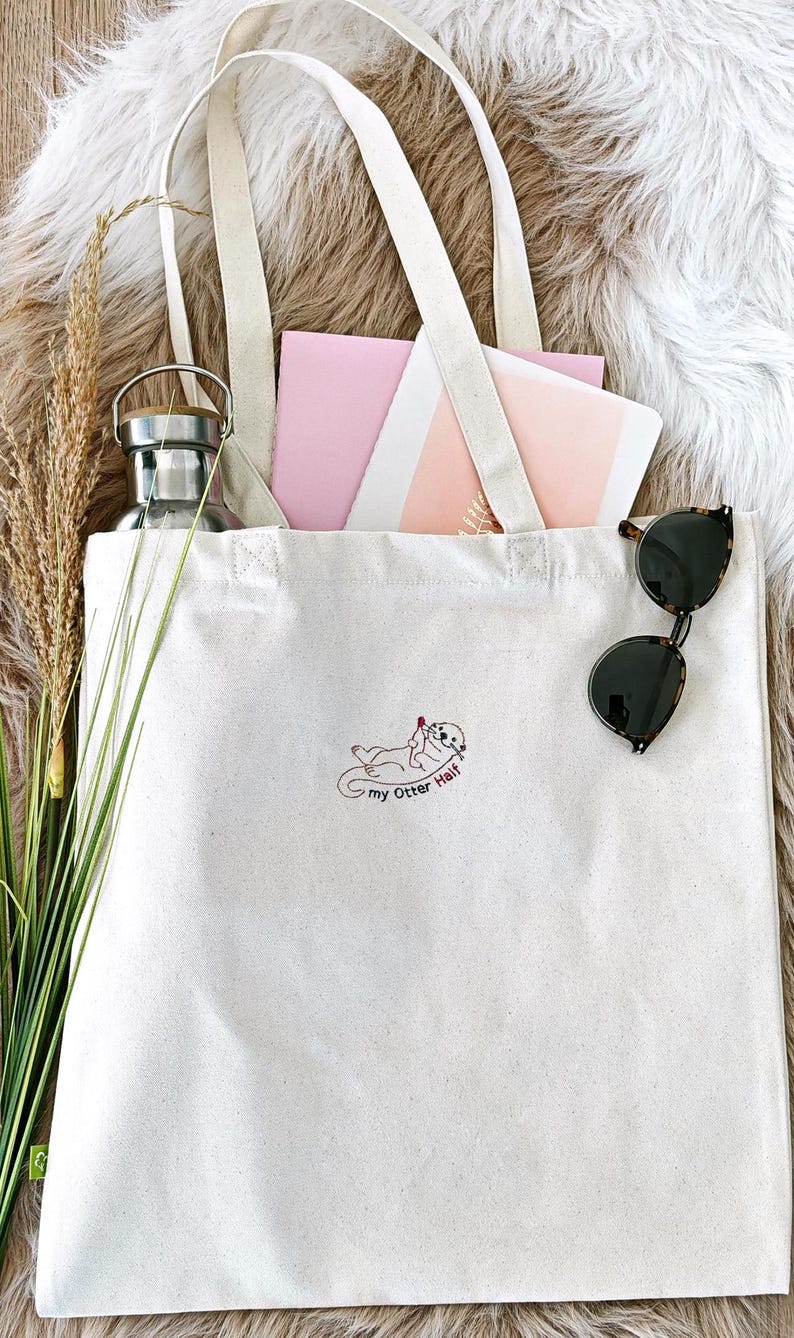 Puede incluir: Un bolso de tela color crema con un bordado de una nutria sosteniendo un coraz&oacute;n rojo y el texto "my Otter Half". El bolso contiene una botella de agua de metal plateado, papeles de color rosa y blanco, y tallos de hierba seca.
