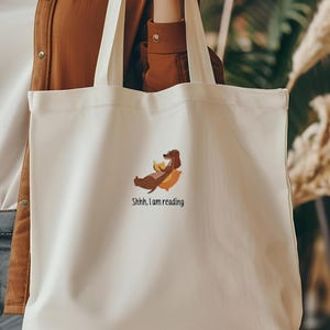 Puede incluir: Una bolsa de tela blanca con una ilustraci&oacute;n de un perro salchicha marr&oacute;n y el texto "Shhh, estoy leyendo".