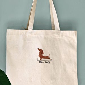 Puede incluir: Una bolsa de lona blanca con un dachshund marr&oacute;n bordado en ella haciendo una pose de yoga. El texto "100% YOGI" est&aacute; bordado debajo del perro.