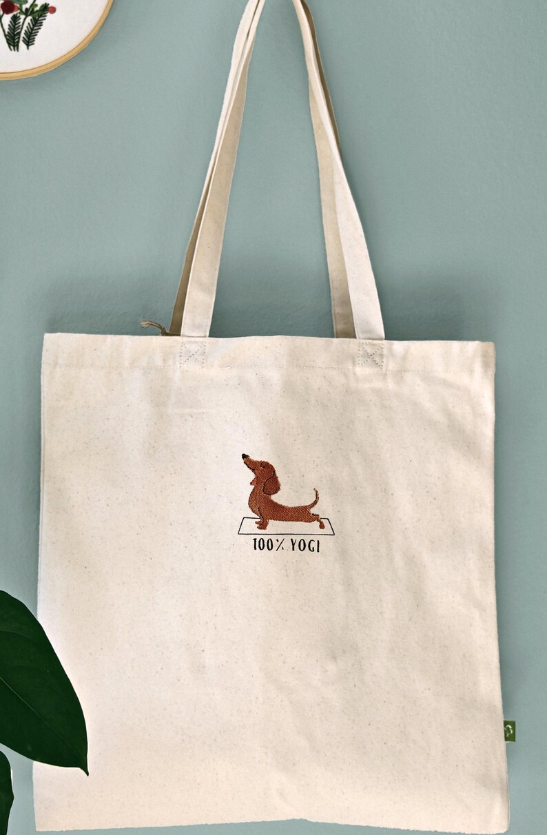 Puede incluir: Una bolsa de lona blanca con un dachshund marr&oacute;n bordado en ella haciendo una pose de yoga. El texto "100% YOGI" est&aacute; bordado debajo del perro.