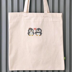 Puede incluir: Una bolsa de tela de color beige claro con un dise&ntilde;o bordado de dos peque&ntilde;os ping&uuml;inos de pie uno al lado del otro. Un ping&uuml;ino lleva un peque&ntilde;o lazo marr&oacute;n, el otro una pajarita negra, y sostienen un coraz&oacute;n rosa entre ambos.